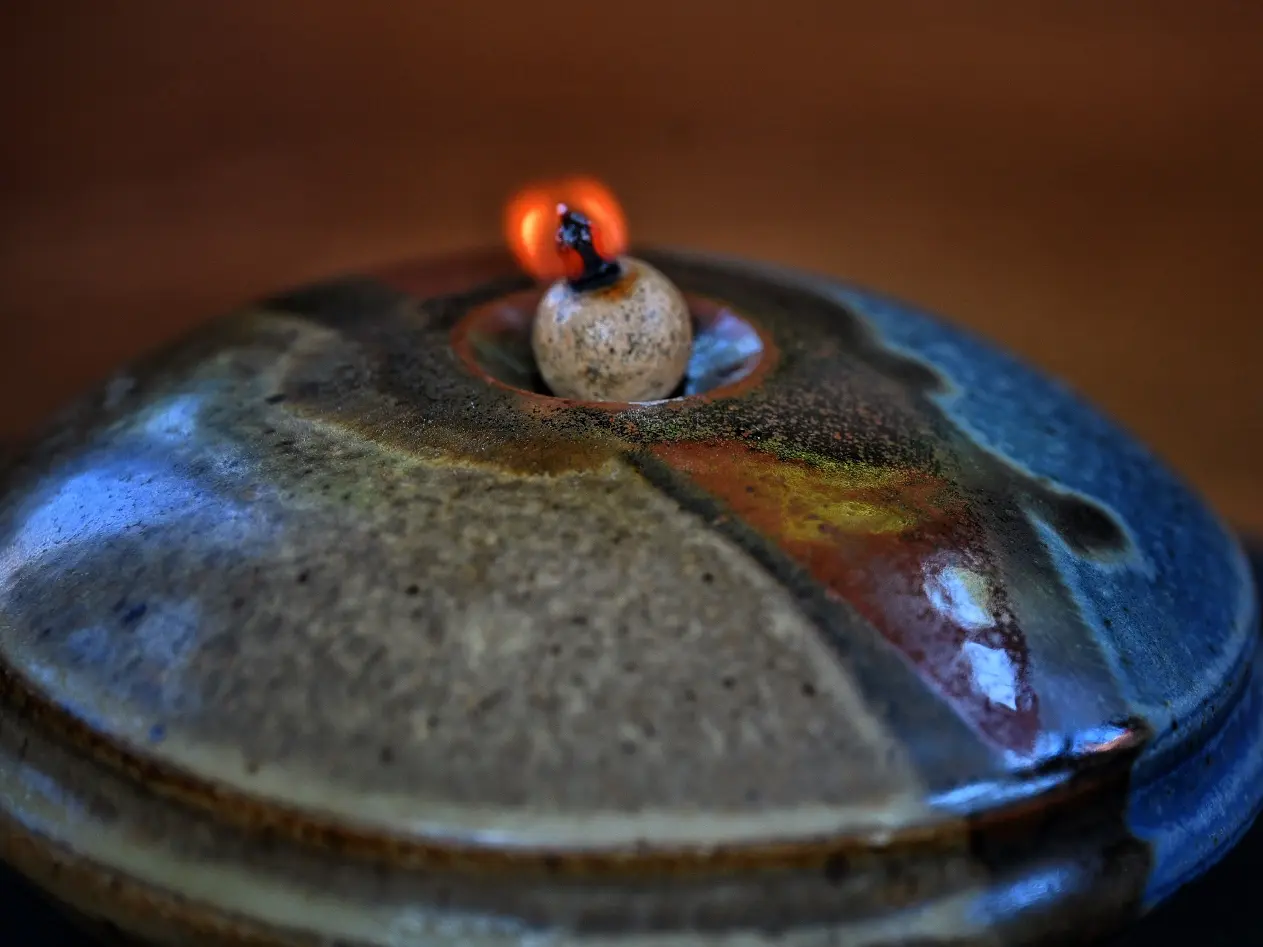 A glowing wick reflected across the blue, red, and earth-toned glaze of an oil lamp