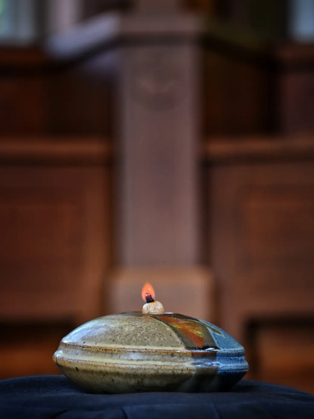 A small flame rises from a ceramic oil lamp resting on a dark cloth